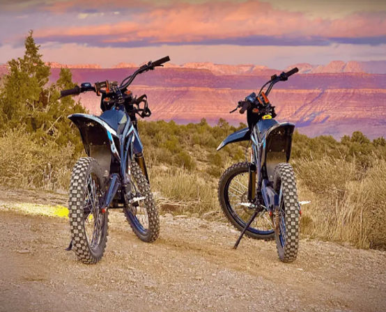 Two off-road electric dirt bikes with knobby tires parked on a desert trail overlooking a red-rock canyon at sunset.