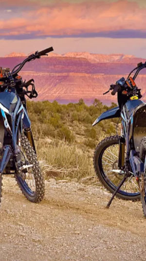 Two off-road electric dirt bikes with knobby tires parked on a desert trail overlooking a red-rock canyon at sunset.