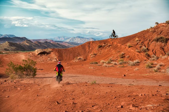 Two dirt bikers on red‑rock desert trails with one riding away and another on a ridge, dramatic mesas and distant mountains under a wide blue sky — off‑road adventure in the American Southwest