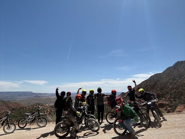 Group of off-road dirt bike riders in colorful helmets posing at a desert canyon overlook with rugged red rock cliffs, motorcycles, and a bright blue sky