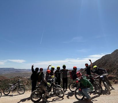 Group of off-road dirt bike riders in colorful helmets posing at a desert canyon overlook with rugged red rock cliffs, motorcycles, and a bright blue sky