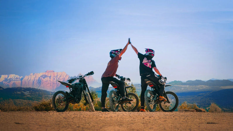 Two helmeted off-road riders high-fiving beside dirt bikes on a dusty overlook, with sunlit red‑rock mesas and a wide desert valley under a clear blue sky