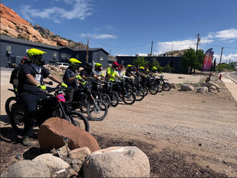 Group ride of electric dirt bikes with riders in neon yellow helmets lined up near red rock cliffs under a bright blue desert sky
