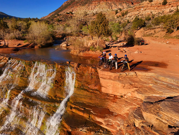 Four helmeted dirt-bike riders paused on a red sandstone cliff beside a cascading waterfall and shallow river in a sunlit southwestern canyon with scrub trees and an ATV in the background.