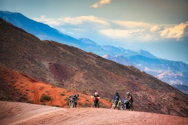 Four dirt bikers on an off-road red-rock desert trail with layered blue mountain ridges and a dramatic cloudy sky — adventurous Southwestern scenery
