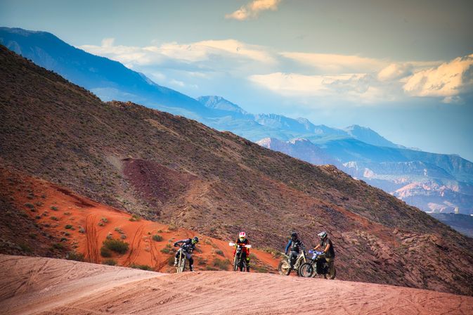 Four dirt bikers on an off-road red-rock desert trail with layered blue mountain ridges and a dramatic cloudy sky — adventurous Southwestern scenery