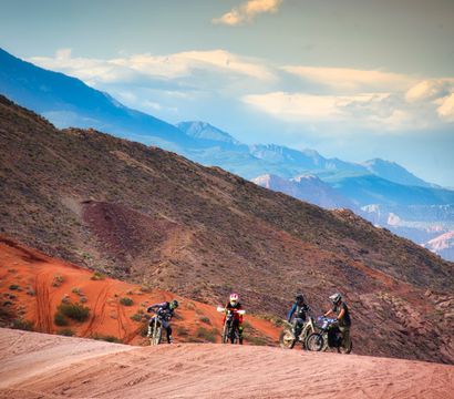 Four dirt bikers on an off-road red-rock desert trail with layered blue mountain ridges and a dramatic cloudy sky — adventurous Southwestern scenery