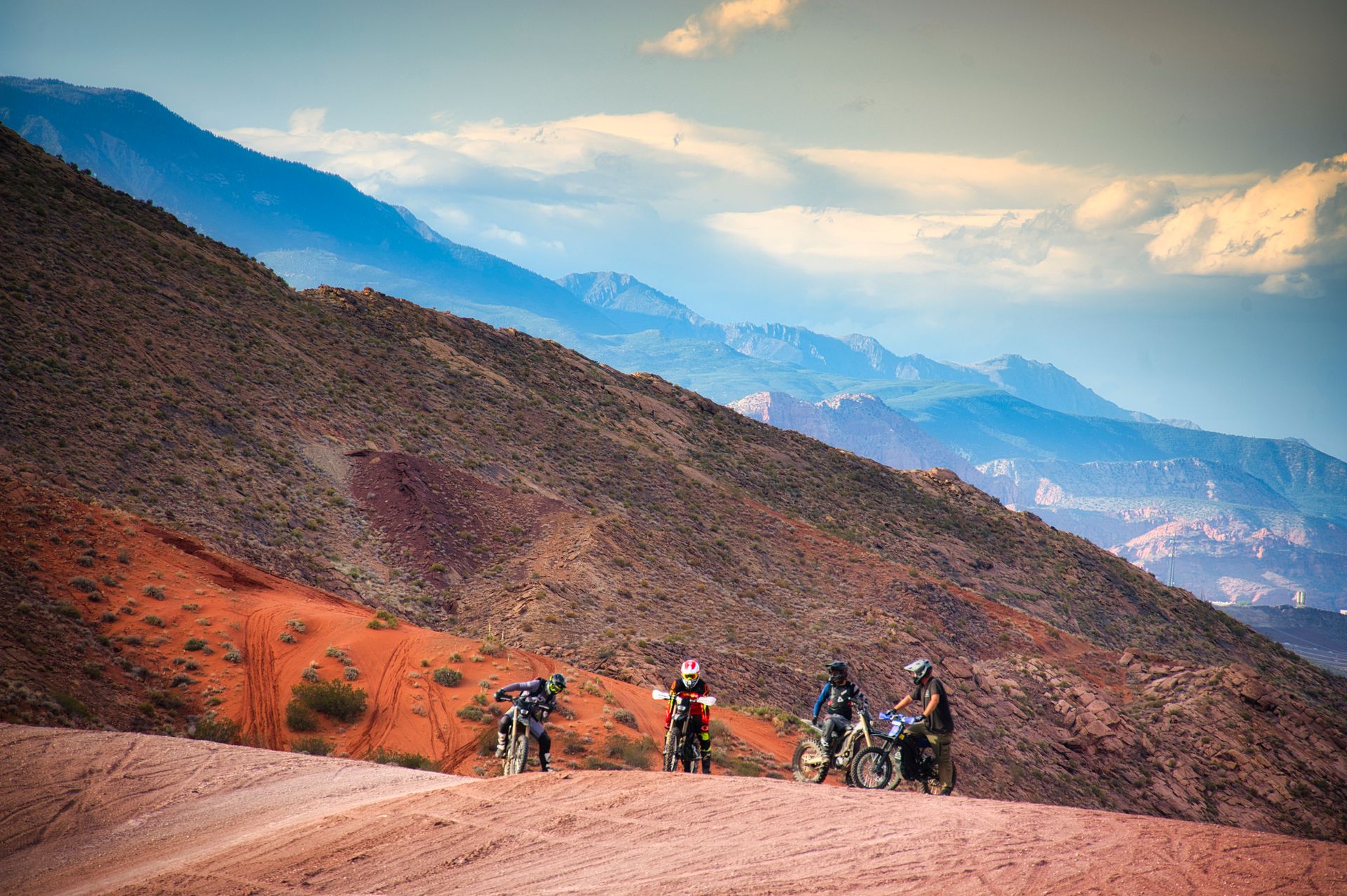 Four dirt bikers on an off-road red-rock desert trail with layered blue mountain ridges and a dramatic cloudy sky — adventurous Southwestern scenery
