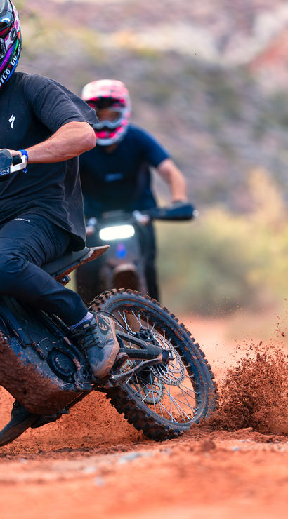 Off-road dirt bike riders on a red-dirt desert trail — helmeted rider leans into a turn, kicking up mud and clay as two riders follow through scrubby terrain