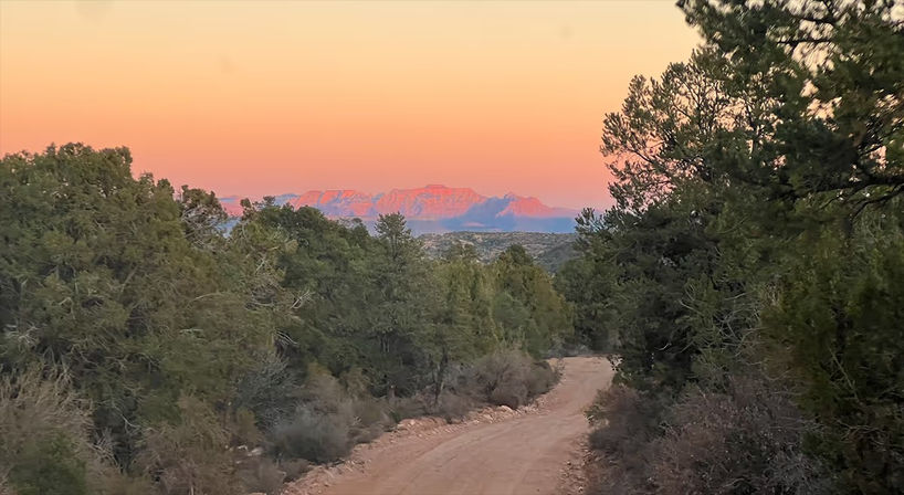 Winding dirt road through evergreen scrub toward distant pink-lit mesas at sunset, pastel orange sky over a scenic southwestern desert landscape.