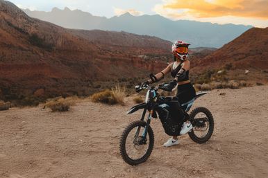 Off-road rider in helmet and goggles on a black dirt bike paused on a red rock desert canyon trail at sunset with layered mountains in the background.