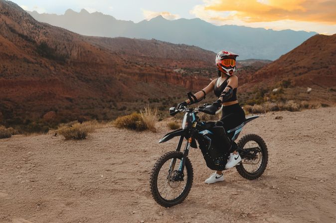 Off-road rider in helmet and goggles on a black dirt bike paused on a red rock desert canyon trail at sunset with layered mountains in the background.