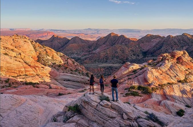 Three hikers stand on layered pink sandstone overlooking sweeping red‑rock canyons and distant mesas in the American Southwest at golden hour — scenic desert hiking overlook.