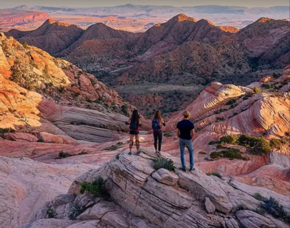 Three hikers stand on layered red-rock sandstone overlooking a sweeping desert canyon at sunset, warm golden light highlighting striped ridges and distant mesas.