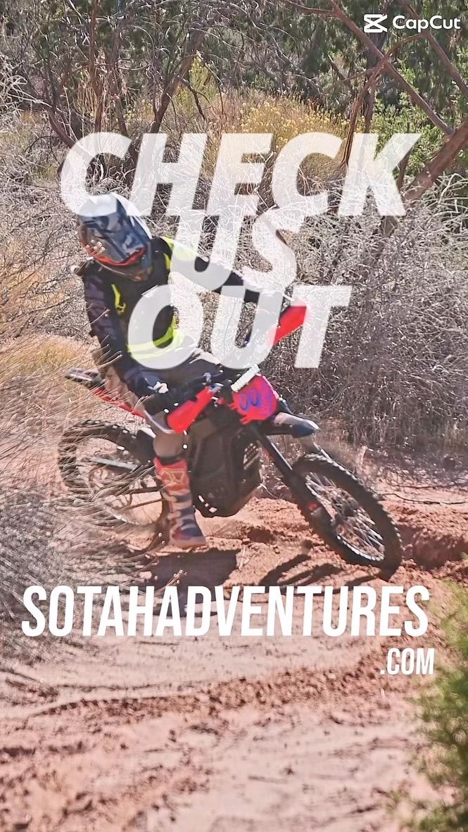 Dirt bike rider in neon gear leaning into a turn on a red sandy desert off-road trail, kicking up dust among scrubby bushes under bright sun — off‑road adventure riding.