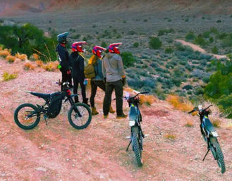 Four helmeted riders with three off-road dirt bikes paused at a red-rock desert overlook, surveying a winding canyon trail.
