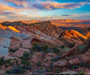 Sunset-lit layered sandstone and red rock formations in a Southwestern desert, dotted with sparse shrubs and distant mesas beneath a colorful sky.