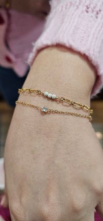 Close-up of a wrist wearing two delicate gold bracelets: a textured link bracelet with three small bead accents and a thin chain with a single bezel-set clear stone, styled with a pink knit sleeve — women's fashion jewelry close-up.