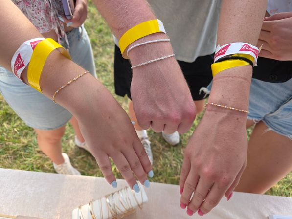 Three outstretched wrists wearing yellow event wristbands and delicate bracelets, painted nails visible, over a table at an outdoor summer festival on grass