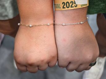 Close-up of two wrists side-by-side wearing delicate matching pearl chain bracelets—one gold, one silver—with a yellow 2023 festival wristband visible on one wrist.