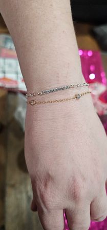 Close-up of a person's wrist and hand wearing two delicate bracelets — a silver pavé bar chain and a thin gold chain with small bezel-set crystals — against a blurred pink and patterned background, wrist jewelry close-up.