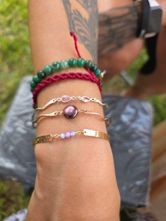 Stacked wrist bracelets on a forearm: green stone bead strand, red macramé cord, gold wavy bangles with pink gems and a purple pearl, blurred grass background