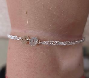 Close-up of a wrist wearing a delicate silver braided chain bracelet with two small round engraved initial discs on soft skin background
