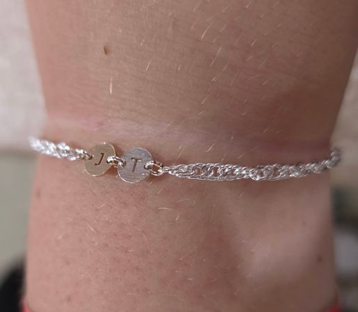 Close-up of a wrist wearing a delicate silver braided chain bracelet with two small round engraved initial discs on soft skin background