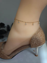 Close-up of an ankle wearing a delicate gold anklet with small dangling crystal charms paired with a glittery nude high-heel pump, fashion jewelry detail.