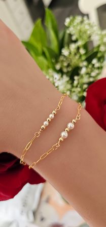 Close-up of a wrist wearing two delicate gold chain bracelets with alternating tiny gold beads and white pearls, photographed over a red rose and white floral background — minimalist fashion jewelry close-up.