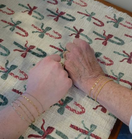 Two hands — one younger and one older — resting side-by-side on a festive Christmas candy-cane patterned tablecloth, both wearing gold bracelets and rings, cozy holiday scene.