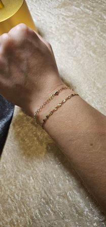 Close-up of a wrist wearing two dainty gold chain bracelets—one beaded link and one floral-link—resting on a soft beige velvet surface, minimalist jewelry styling.