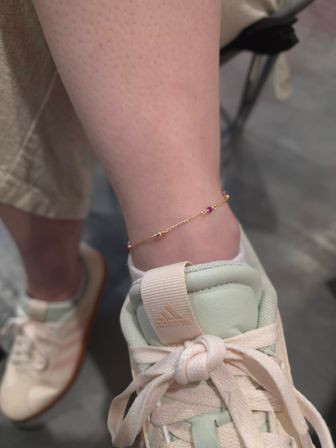 Close-up of an ankle wearing a delicate gold chain anklet with a small pink bead, resting on a mint-and-cream pastel sneaker with knotted laces — casual street style.