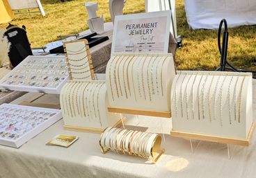 Sunlit outdoor artisan market jewelry display featuring a 'Permanent Jewelry' sign and rows of delicate gold and silver bracelets and anklets on white stands at a vendor booth.