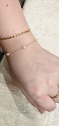 Close-up of a light-skinned wrist and hand wearing two stacked gold bracelets — a chunky curb chain and a delicate chain dotted with small pearls — plus a simple gold ring on a beige velvet background.