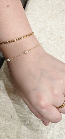 Close-up of a light-skinned wrist and hand wearing two stacked gold bracelets — a chunky curb chain and a delicate chain dotted with small pearls — plus a simple gold ring on a beige velvet background.