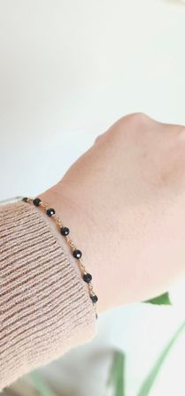 Close-up of a wrist wearing a delicate gold chain bracelet with small faceted black beads, resting over a beige ribbed sweater cuff — minimalist jewelry shot.