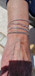 Close-up of a sunlit wrist and hand outdoors wearing four delicate sparkling silver chain bracelets on textured skin with light sun spots.