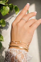 Close-up of a hand with French manicure wearing a thin diamond band and stacked gold bracelets — hammered bangle, pearl chain and link bracelet — over a lace sleeve in warm sunlight with green plant leaves in the background.