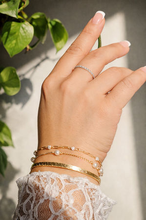 Close-up of a hand with French manicure wearing a thin diamond band and stacked gold bracelets — hammered bangle, pearl chain and link bracelet — over a lace sleeve in warm sunlight with green plant leaves in the background.