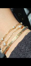 Sunlit wrist wearing three sparkling gold bracelets — textured gold bangle, delicate pearl chain and beaded link chain, photographed inside a car.
