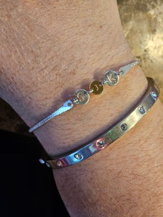 Close-up of a wrist wearing two silver bracelets: a delicate chain with three round initial charms reading S, J, K and a shiny rhinestone-studded bangle.