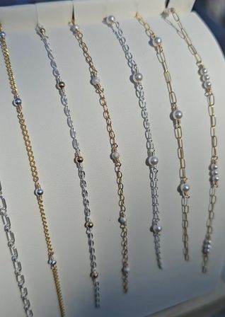 Close-up of delicate gold and silver chain bracelets with tiny pearls and metallic bead accents displayed vertically on a white jewelry stand — minimalist fashion accessory close-up
