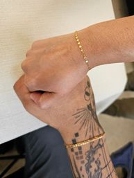 Close-up of two wrists wearing matching delicate gold chain bracelets, one wrist with geometric and dotwork tattoos, hands touching over a light textured background