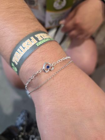 Close-up of a wrist wearing a green festival wristband and delicate silver double-chain bracelet with a heart-shaped charm set with a small blue stone.
