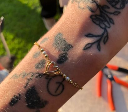 Close-up of a tattooed forearm wearing a gold heart charm bracelet with tiny pearls, sunlit backyard grass and red-handled pliers blurred in the background.