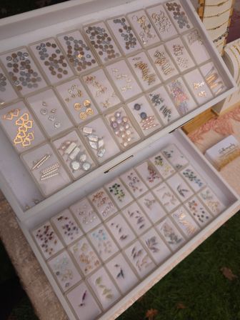 White two-tier jewelry display tray filled with organized earring studs, charms, pearls and colorful gemstone components on an outdoor market table.