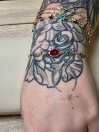 Tattooed wrist with black floral design wearing delicate gold chain bracelets centered on an oval red gemstone with small blue and pink stones and a tiny star tattoo on the hand