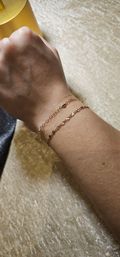 Close-up of a wrist wearing two dainty gold chain bracelets—one beaded link and one floral-link—resting on a soft beige velvet surface, minimalist jewelry styling.