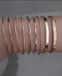 Close-up of a wrist wearing a stacked set of slim gold bangles with mixed textures — smooth, twisted, wavy, hammered and floral-engraved — minimalist boho jewelry accessory.